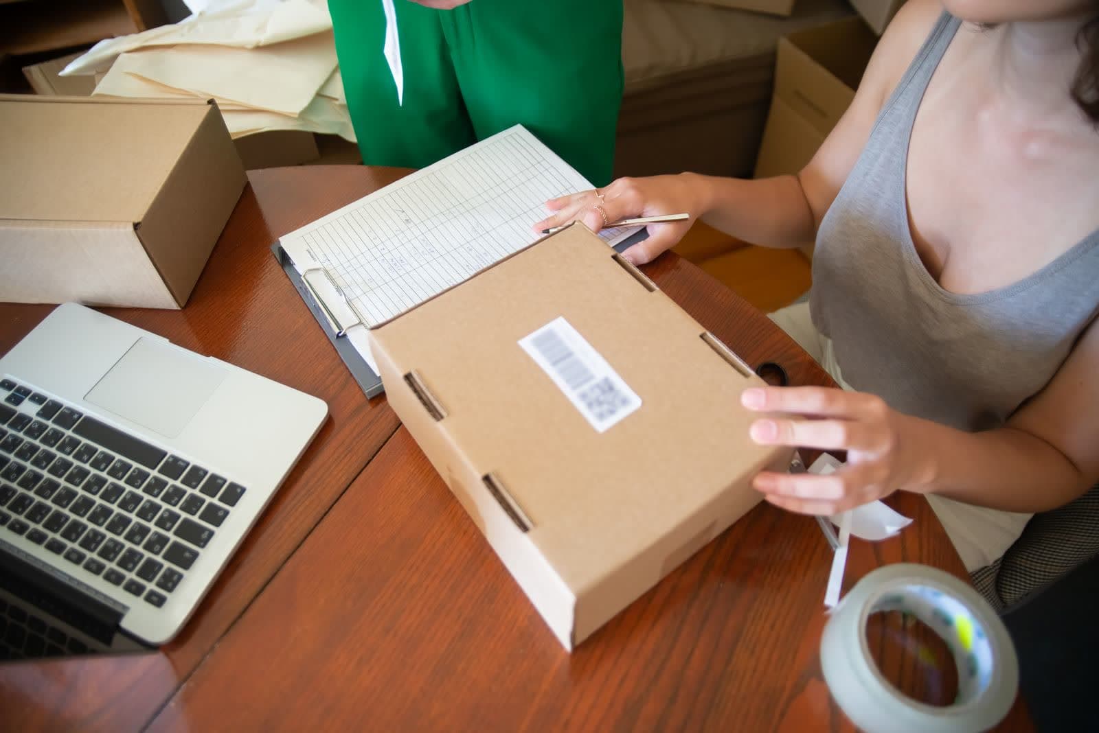 Hands taping a cardboard parcel closed beside an open laptop